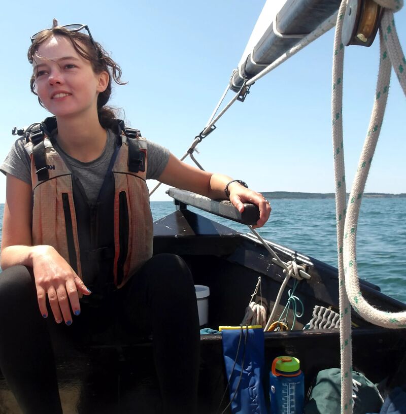 A young woman is sailing on a sunny day. She is wearing a life vest and dark clothing. She is holding the tiller of the sailboat, looking off to the side. The boat is on the water, and the sky is clear. There is a mast and ropes visible, and the water is calm.